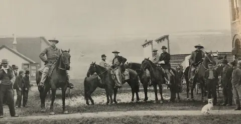 A historic black and white photograph of men on horseback and on foot outdoors.