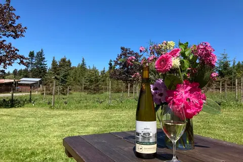 A bottle of white wine, a glass, and a bouquet of flowers sit on a wooden table outdoors.