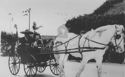 A historic black and white photo of people riding in a horse-drawn carriage.
