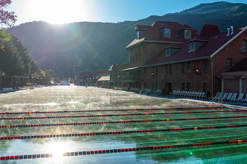 A large outdoor swimming pool with steam rising from the water, flanked by buildings and mountains.