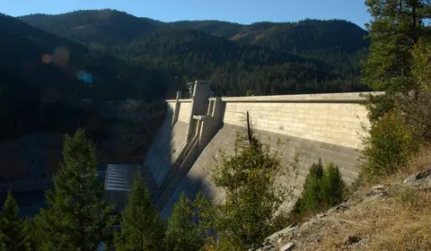 A large concrete dam stands before forested hills and mountains.