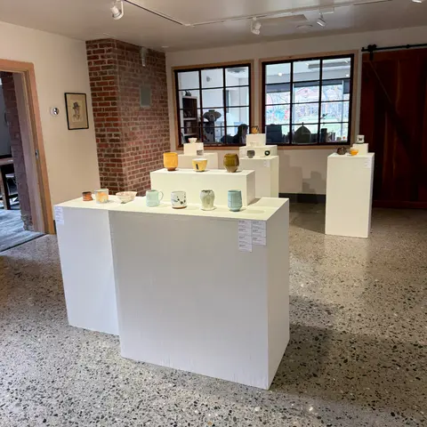 A gallery interior displays ceramic mugs and bowls on white pedestals.