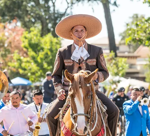 A man in a charro outfit rides a horse in a parade with musicians.