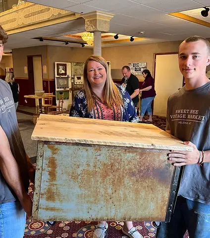 Three people pose while two young men hold a rustic metal cart with a wooden top indoors.