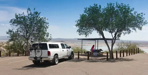 A pickup truck with a bicycle rack is parked near a shaded picnic table overlooking a body of water.