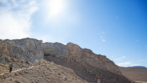 A person hiking near a rocky cliffside under a bright blue sky.