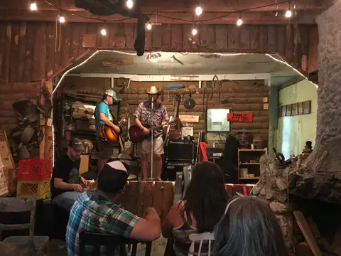 A small band performs on a stage inside a rustic log building with an audience watching.