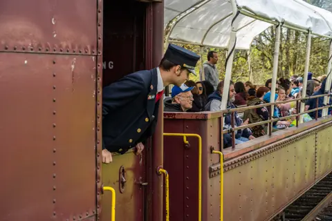 A train conductor looks out the doorway of a train car carrying passengers.