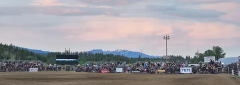 A wide shot of a rodeo arena with crowds watching under a cloudy sky.