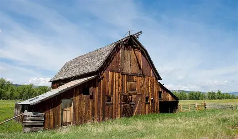 A rustic wooden barn stands in a grassy field under a blue sky.