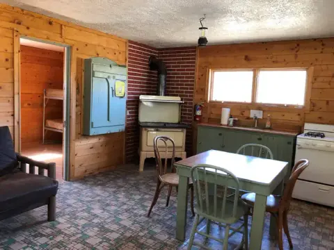 The interior of a rustic cabin featuring a kitchen, dining table, wood stove, and a doorway leading to a bedroom with bunk beds.