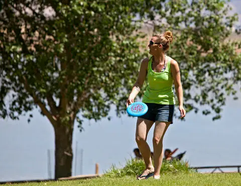 A woman holds a flying disc outdoors near a tree and water.