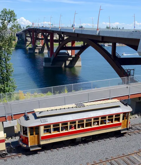 A vintage trolley car travels on tracks near a large bridge over water.