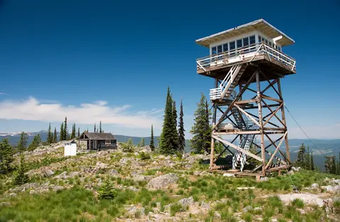 A wooden fire lookout tower stands on a grassy mountain summit with a cabin and trees.