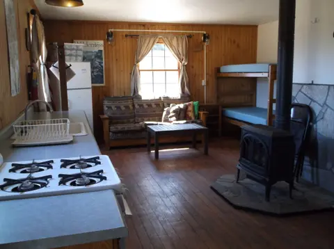 Interior view of a rustic cabin featuring a kitchen counter, living area, wood stove, and bunk beds.