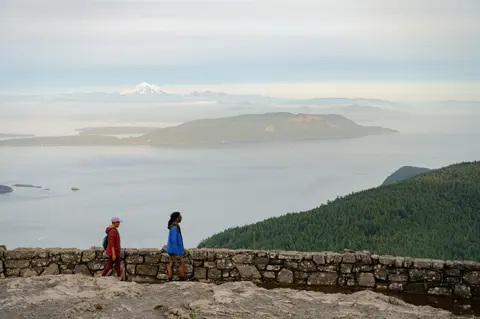 Two hikers walking along a stone wall overlooking a scenic landscape with water, islands, and distant mountains.