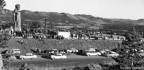 Historic black and white photograph showing a large crowd gathered near a monument with cars parked below.