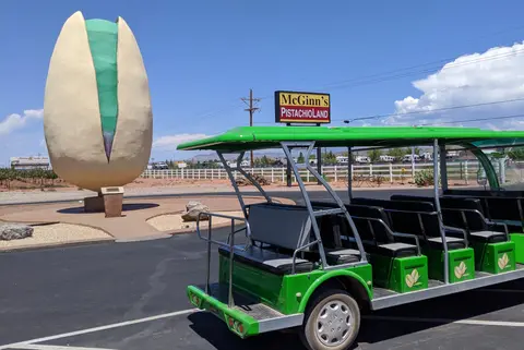 A large pistachio sculpture stands next to a green tour vehicle in an outdoor parking area.
