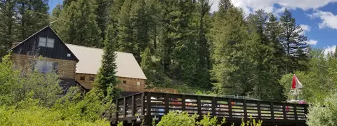 A rustic lodge sits in front of a dense pine forest with a wooden deck and pool area in the foreground.