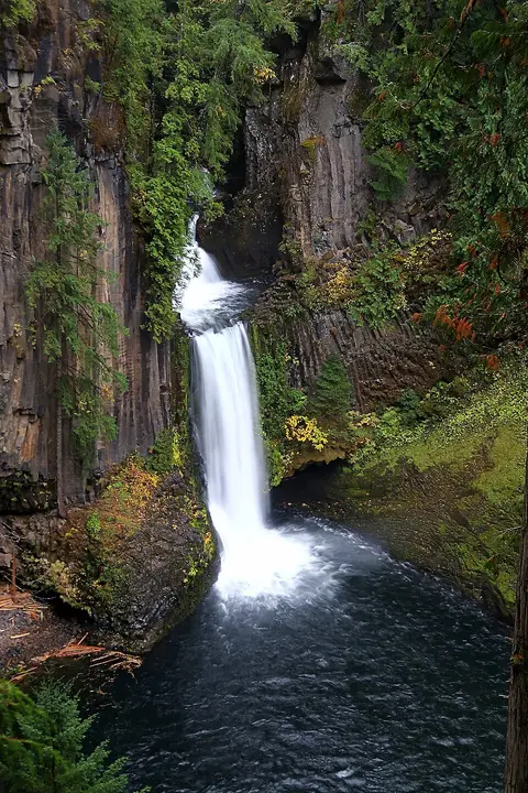 A waterfall plunges into a dark pool surrounded by rocky cliffs and green trees.