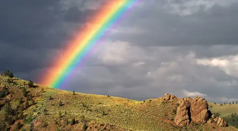 A bright rainbow arches over a grassy hill and rocky outcropping under stormy skies.