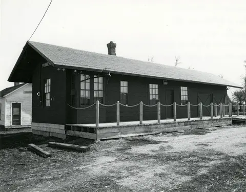 Black and white exterior photo of a historic single-story wooden building with a long platform and rope railing.