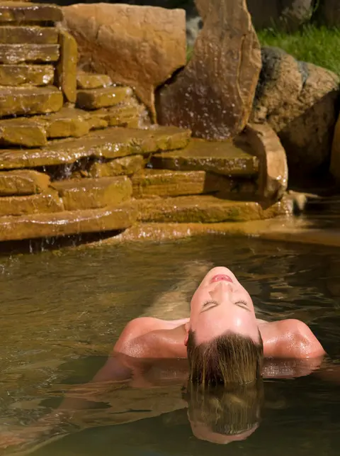A woman relaxing and floating in a natural hot spring or pool with a stone waterfall background.