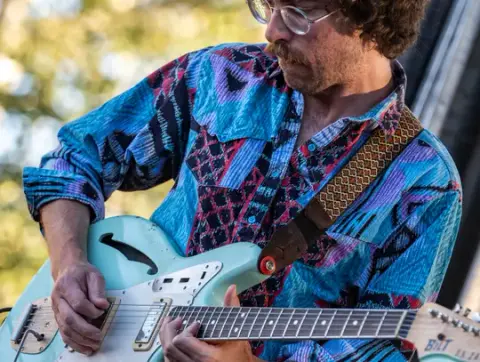 A man plays an electric guitar while wearing a patterned shirt and glasses.