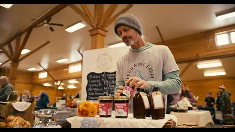 A vendor stands behind a table with jars of goods inside a wooden building.