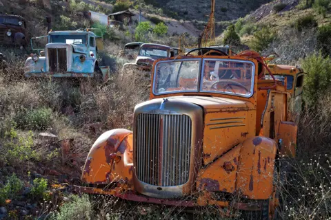 Old rusted vintage trucks sit abandoned on a hillside overgrown with dry grass and brush.