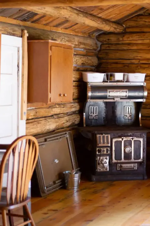 A rustic interior shows a wooden cabinet and an antique stove in a log cabin.