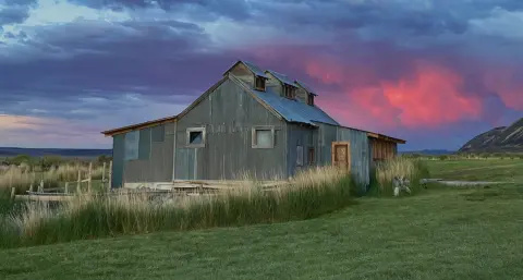 A rustic metal building stands in a grassy field beneath a colorful sunset sky.