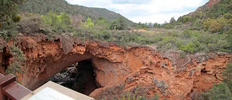 A large natural rock bridge spans a canyon surrounded by green hills and trees.