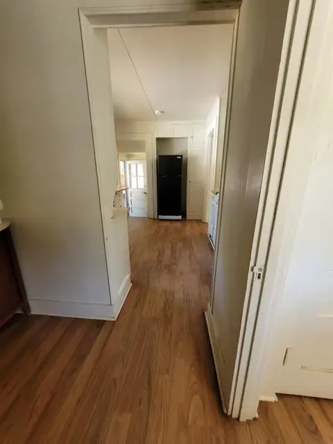 A view through a doorway into a hallway with wooden floors and a black refrigerator at the end.