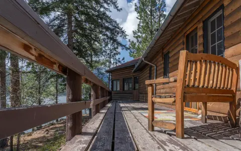 A wooden bench sits on the porch of a rustic log cabin overlooking trees and water.
