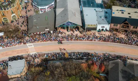 An aerial view shows a large crowd gathered along a street lined with buildings.