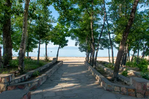 A stone pathway leads through trees toward a sandy beach and water.