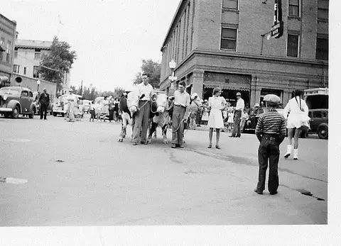 A black and white photograph shows people leading oxen on a town street.