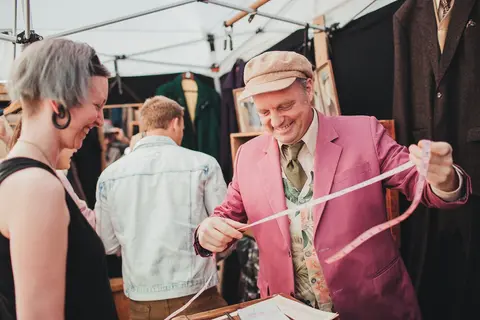 A tailor holds a measuring tape while smiling at a customer at a market stall.