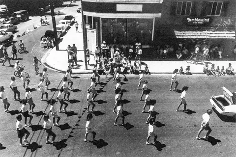 An elevated black-and-white photograph shows people marching down a street during a community event.