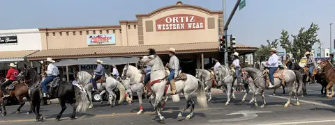 A street parade featuring people riding horses past storefronts.