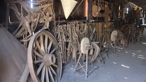 An interior view of a rustic barn filled with vintage farm equipment, wooden wagon wheels, and hanging harnesses.