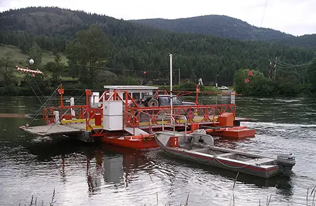 A ferry boat carrying a vehicle across a river with a small motorboat alongside.