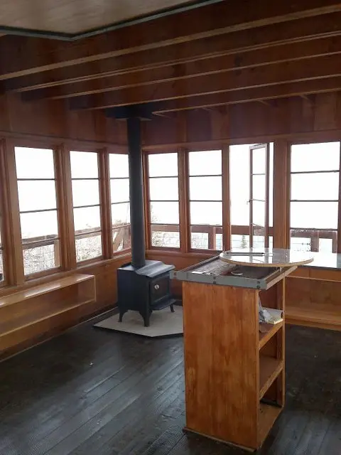 An interior view of a wooden cabin with many windows, a wood-burning stove, and a table.