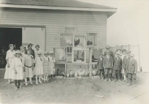 A group of children posing with stacked cages of chickens outside a building.