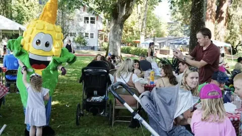 A costumed corn mascot interacts with children and families at an outdoor gathering.