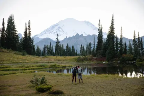Two hikers stand by a reflective pond looking at a snow-capped mountain.