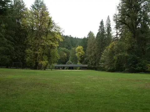A large grassy field surrounded by dense forest with a picnic shelter in the distance.