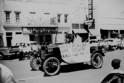 A historic black and white photograph of an old car driving down a street.