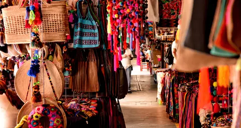 A market stall displays vibrant woven bags, colorful textiles, and hanging pom-pom decorations along a corridor.
